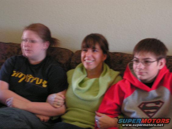 Three people, two women and a boy, sit on a couch. The woman on the left wears a black shirt with yellow text. The middle woman wears a green cowl neck sweater. The boy on the right wears a red and gray Superman hoodie