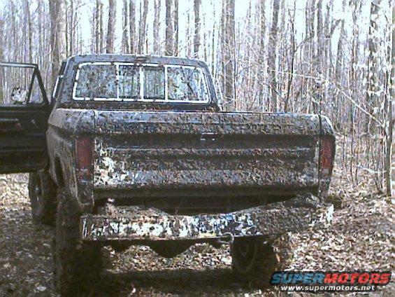 Mud-covered pickup truck rear view, driver's door open, in a wooded area