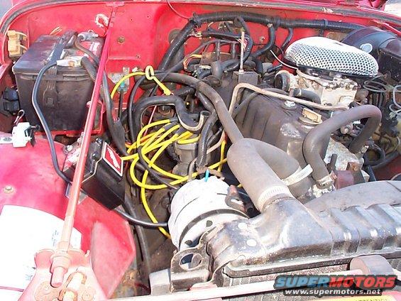 Engine bay of a red Jeep, showing the battery, carburetor, air filter, and wiring