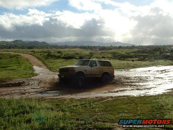 1995 Ford Bronco driving through a muddy field with hills in the background