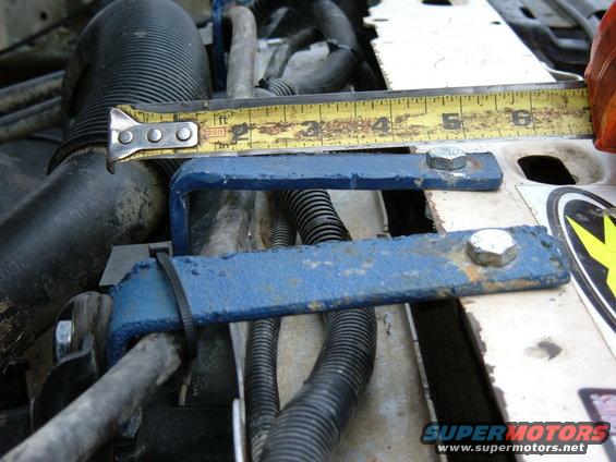 Engine bay with blue metal brackets and a tape measure. Black hoses and wiring are visible