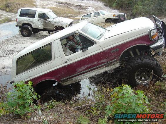 White and maroon 1993 Ford Bronco stuck in mud, front right wheel deep in water
