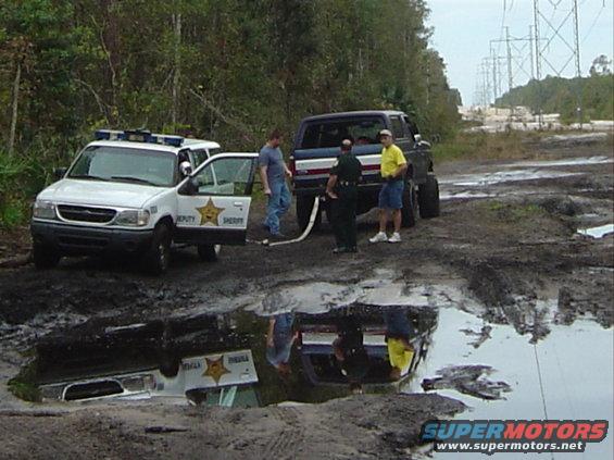 Ford Explorer police SUV and a lifted Ford Bronco stuck in mud, with people standing nearby