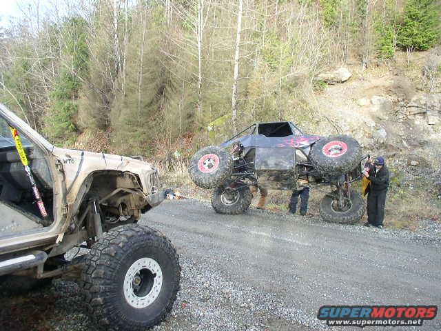 Off-road buggy with flames on the side, and a Jeep in the foreground. People are working on the buggy