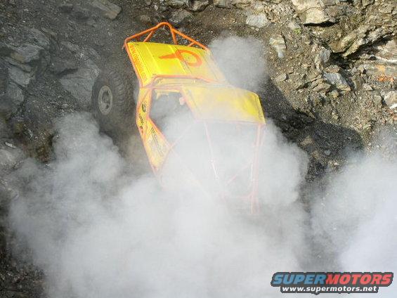 Yellow off-road vehicle with orange roll cage climbing rocky terrain, kicking up smoke