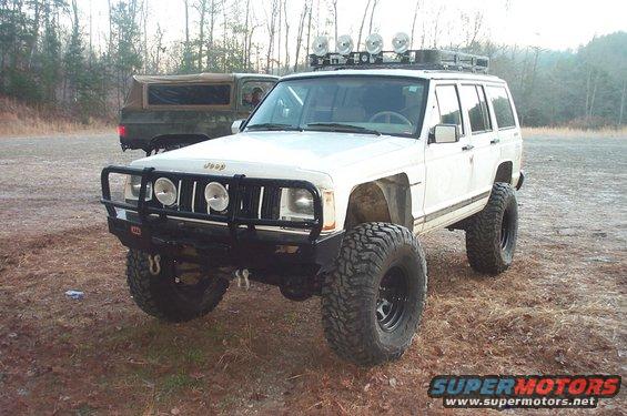 White Jeep Cherokee XJ with aftermarket bumper and large tires. Another SUV is parked behind it