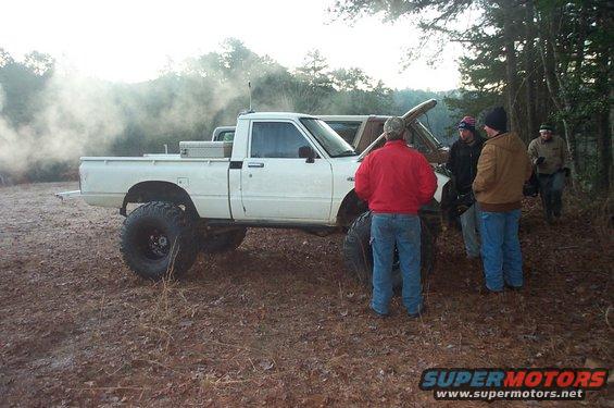 White pickup truck with oversized tires and a toolbox in the bed, parked on a dirt road with trees in the background