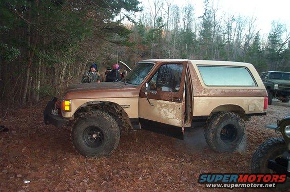 1987 Ford Bronco with lifted suspension and two-tone paint, parked in a wooded area with people around