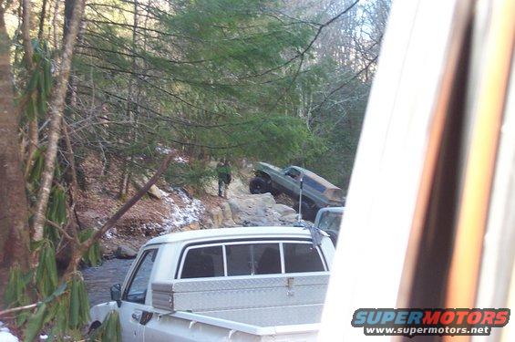Two off-road vehicles navigate a rocky, wooded trail with snow patches