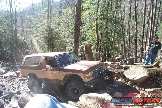 Brown and tan SUV on a rocky trail with trees around. A person stands to the right