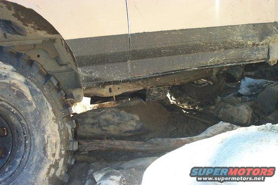 Off-road vehicle chassis and tire stuck on rocks. The vehicle is light-colored with a black trim panel along the lower body. The tire is large and knobby