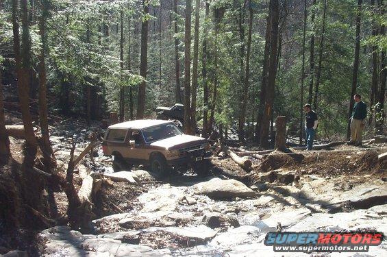 1987 Ford Bronco navigating a rocky stream bed in a forest. Another truck is visible in the background