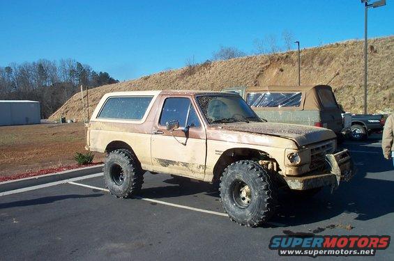 Tan and brown Ford Bronco with large off-road tires, dented front bumper, and visible rust