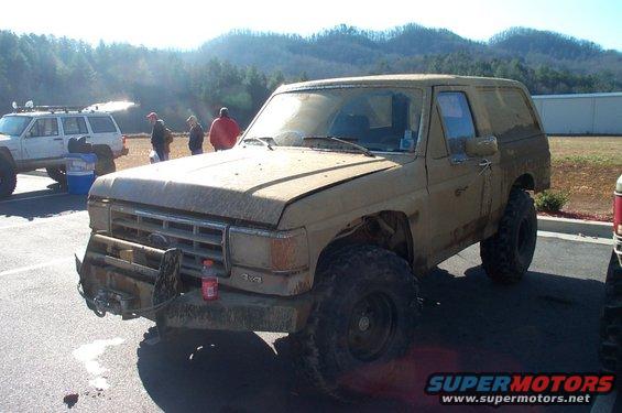 1987 Ford Bronco with aftermarket bumper and winch, covered in mud. Another SUV is parked to the left