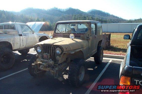 Tan Jeep CJ-5 with a winch and large tires parked next to a white pickup truck