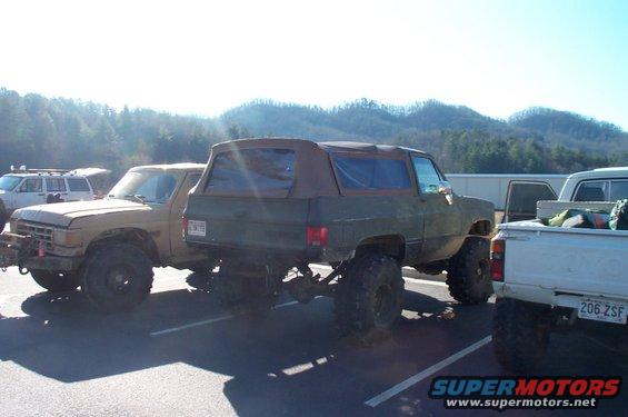 Several lifted off-road vehicles parked in a lot, including a tan pickup and a green SUV with a soft top