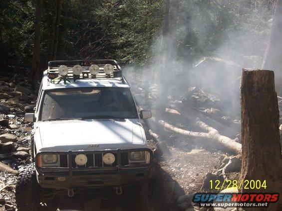White Jeep Cherokee XJ with roof rack and off-road lights driving on a rocky trail with smoke