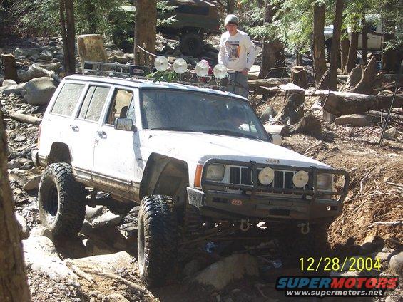 White Jeep Cherokee on rocks with a person standing behind it. Another vehicle is visible in the background