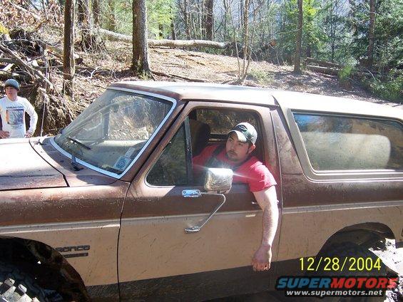 Man in red shirt leans out window of brown Ford Bronco on muddy trail. Another person stands by the front fender