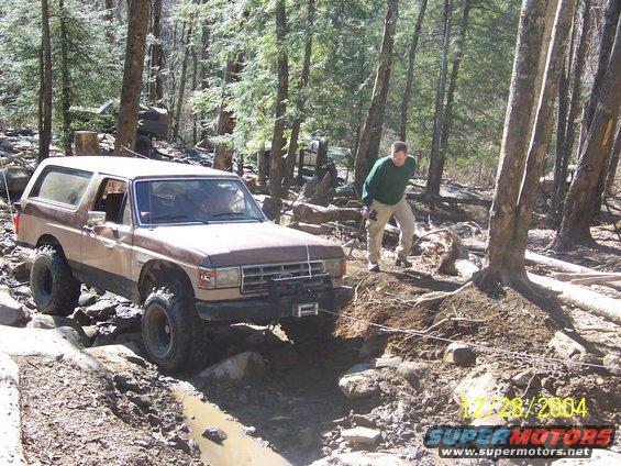1987 Ford Bronco navigating a muddy, rocky trail with a man standing nearby