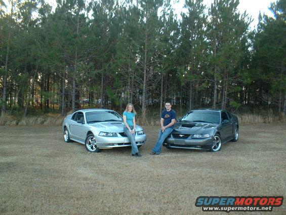 Silver and gray Mustangs parked outside with a man and woman leaning on them