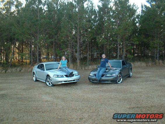 Two 2000 Ford Mustangs, one silver and one gray, parked outdoors with people sitting on hoods