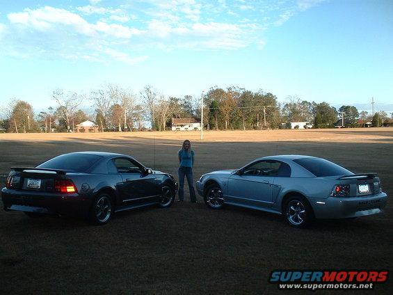 Two 1999 Ford Mustangs, one dark gray and one silver, parked on grass with a woman standing between them