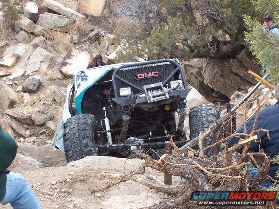 Modified off-road vehicle with GMC grille on rocky trail. Large tires and roll cage are visible