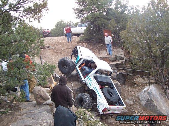 White off-road vehicle with large tires climbing rocks, teal accents, spare tire in bed