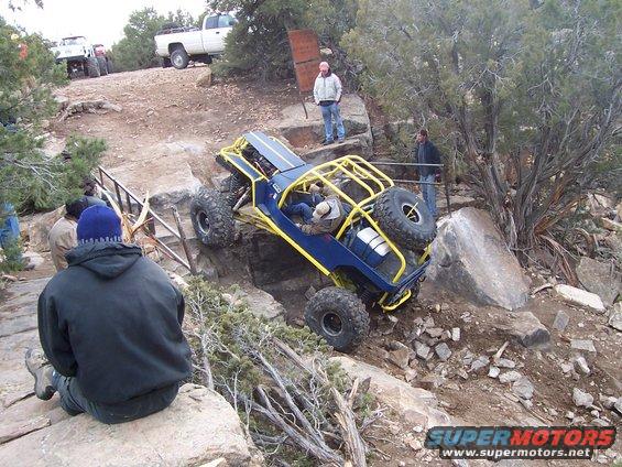 Blue buggy with yellow roll cage climbing rocks. White pickup truck in background