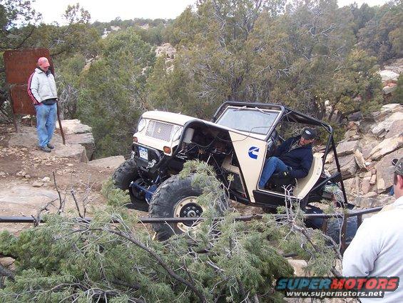 Off-road buggy with large tires navigating rocky terrain. Driver is in the seat, and a person stands nearby