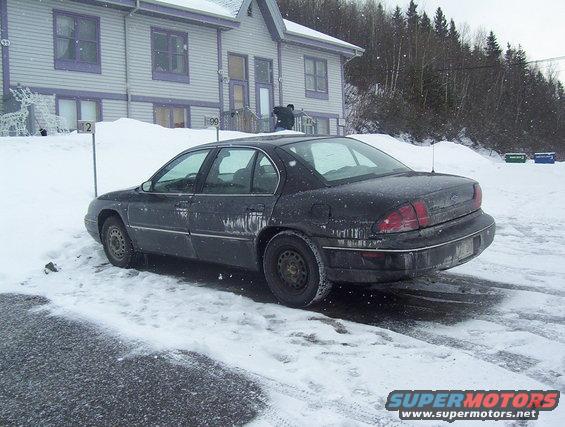 Black sedan parked in snow, rear 3/4 view. Water streaks on body
