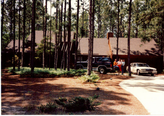 Dark blue SUV and a white sedan parked in front of a house in a wooded area. Three people stand near the SUV