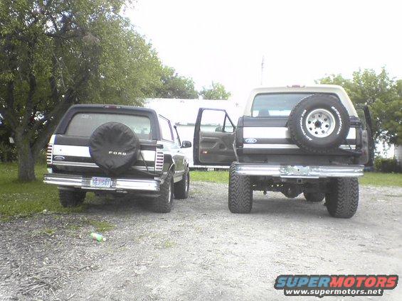 Two Ford Broncos parked side-by-side, both showing their rear ends. The one on the left has a spare tire cover with 'FORD' on it. The one on the right has its spare tire mounted externally