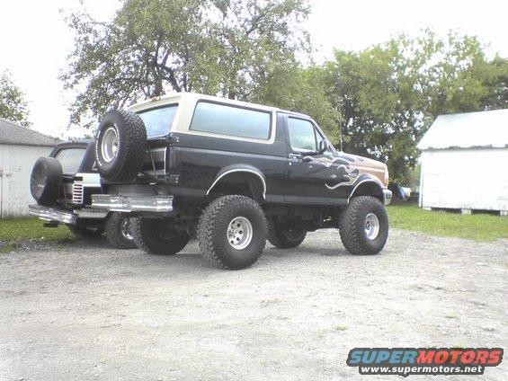 Black and tan lifted Ford Bronco with flame decals and spare tire on rear bumper, parked on gravel