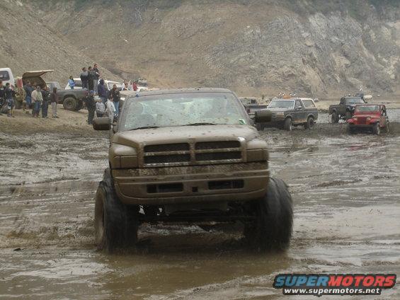 Muddy Dodge Ram pickup truck with large tires driving through a muddy area. Other trucks and people are visible in the background