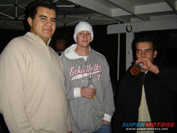 Three men stand together outdoors at night, one drinks from a bottle