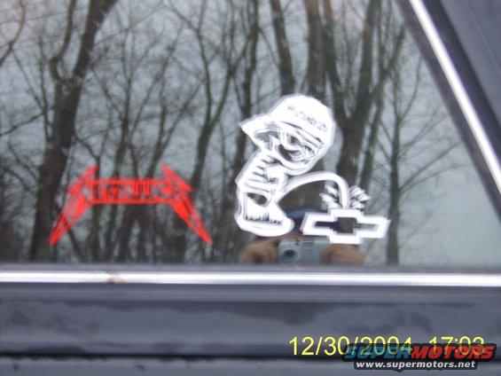 Car window with Metallica logo sticker, a cartoon character sticker, and a Chevrolet logo sticker. Reflection shows trees and a camera
