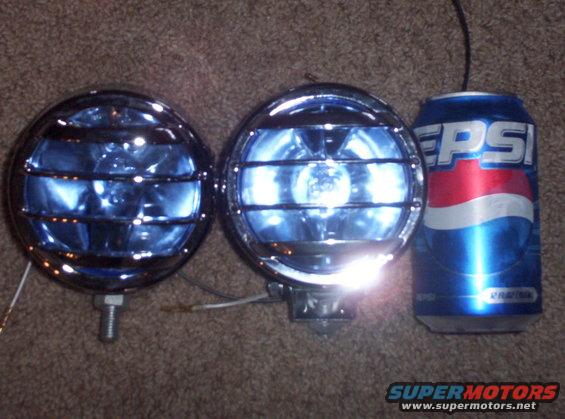 Two chrome-bezeled fog lights with blue lenses and protective grilles sit next to a Pepsi can