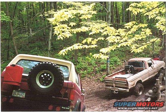 Red SUV's rear with spare tire and gas can, next to a beige pickup truck on a muddy trail