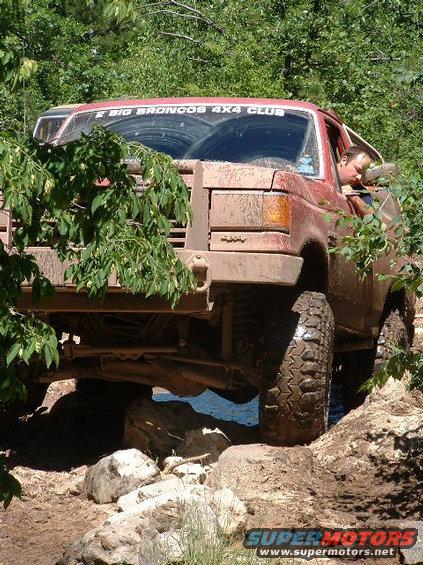 Mud-covered red 1987 Ford Bronco navigating rocky terrain, front 3/4 view