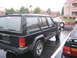 1991 Ford Bronco - A black Jeep Cherokee is seen from the rear quarter angle in a parking lot on a rainy day, with other vehicles and buildings in the backgro…