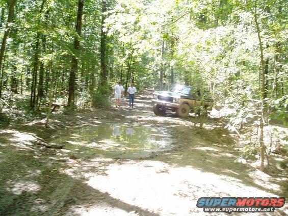 1978 Ford Bronco driving on a muddy, wooded trail with two people walking nearby