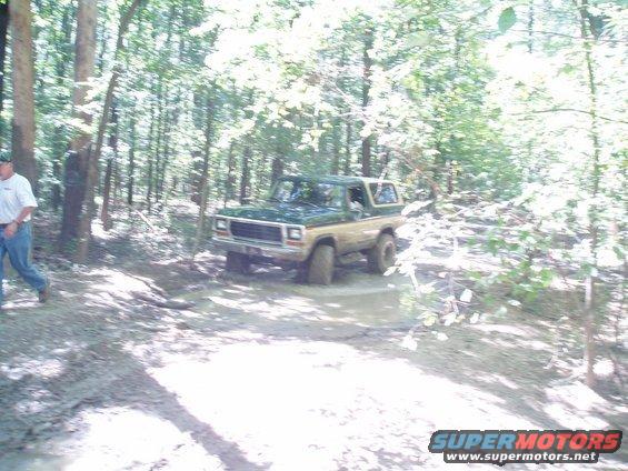 Ford Bronco driving through mud in a wooded area. A person walks nearby