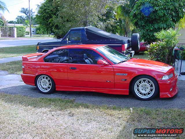 Red 1995 BMW M3 coupe, side view, with a blue truck behind it