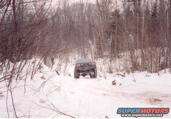 Dark SUV driving on snowy forest road. Appears to be a 1990s Ford Bronco