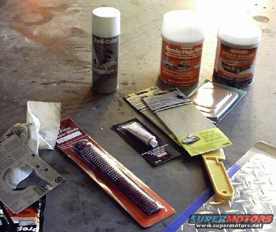 Spray paint, body filler, sandpaper, and a body file are laid out on a concrete floor
