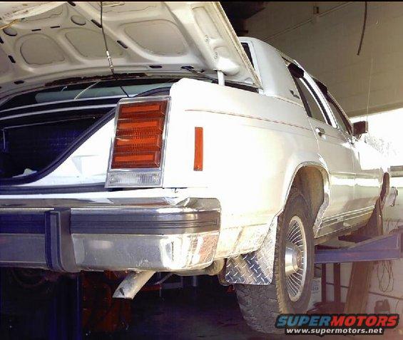 White sedan on a lift, rear view. Open trunk, chrome bumper, diamond plate mud flap