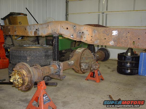 Truck frame and front/rear axles supported by jack stands in a garage. A John Deere mower is visible behind the frame