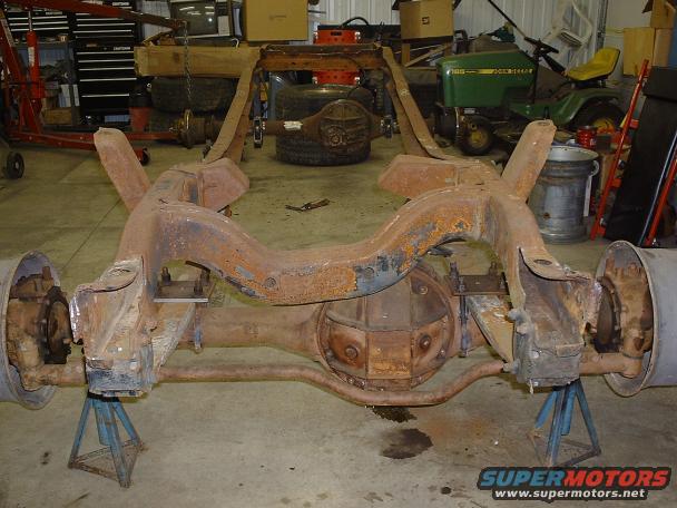 Rusty truck frame and rear axle assembly on jack stands in a garage. A John Deere mower is in the background